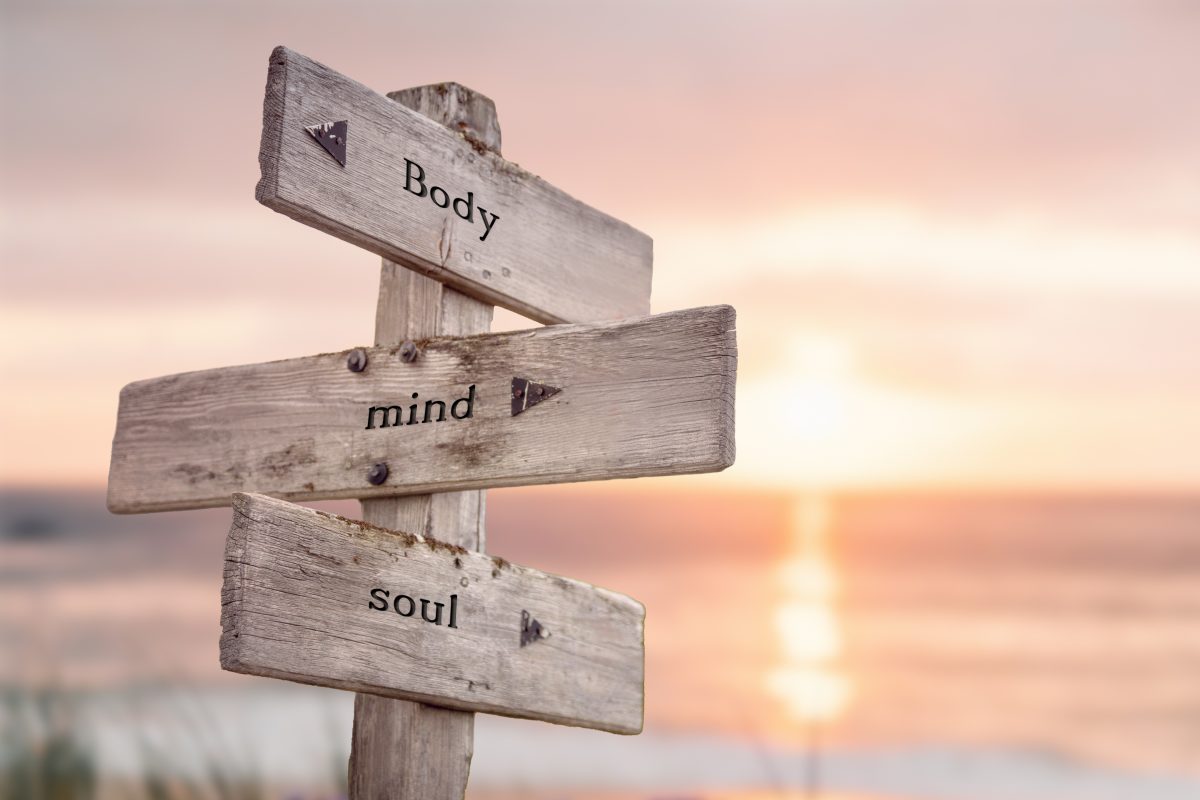 Wooden street sign reading mind, body, and soul with sunset and water in the background, symbolizing a spiritual journey.